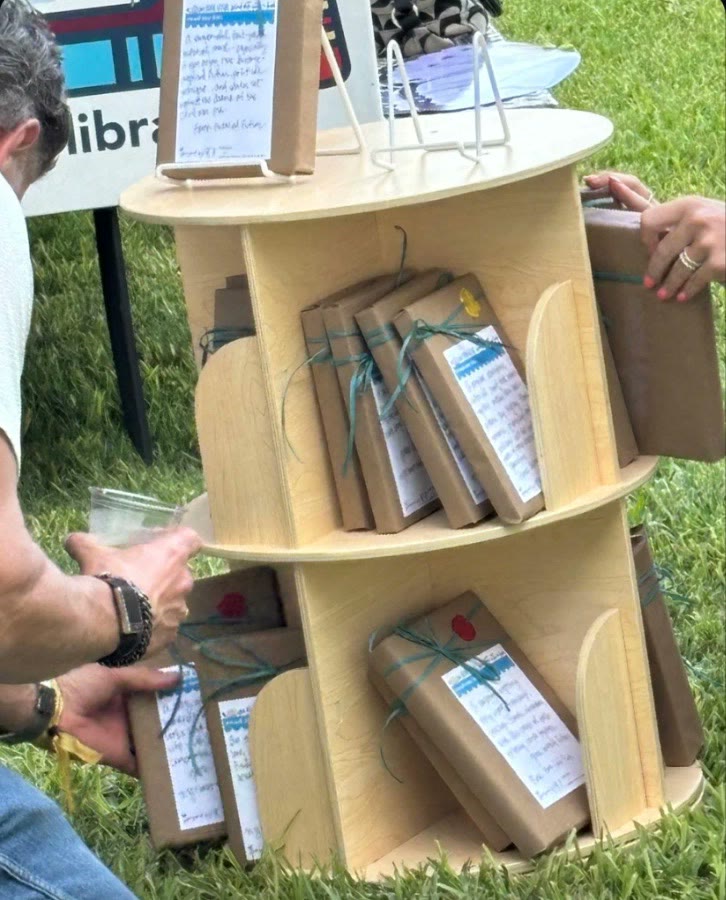 Books wrapped in brown paper and tied with ribbon, each with a handwritten note, on a round wooden shelf on the grass