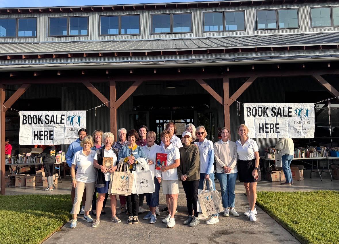 Friends volunteers in white Friends polo shirts standing under two Book Sale Here banners at Caliterra, with tables and boxes of books behind them