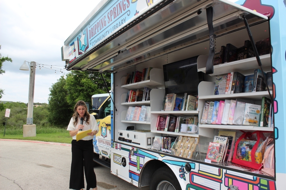 The Dripping Springs Community Library's mobile library van, open and stocked with books, with a woman speaking into a microphone beside it