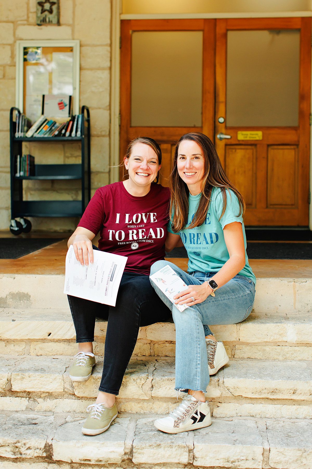 Two smiling women in 'I love to read' shirts sitting on the stone steps outside the library's wooden doors