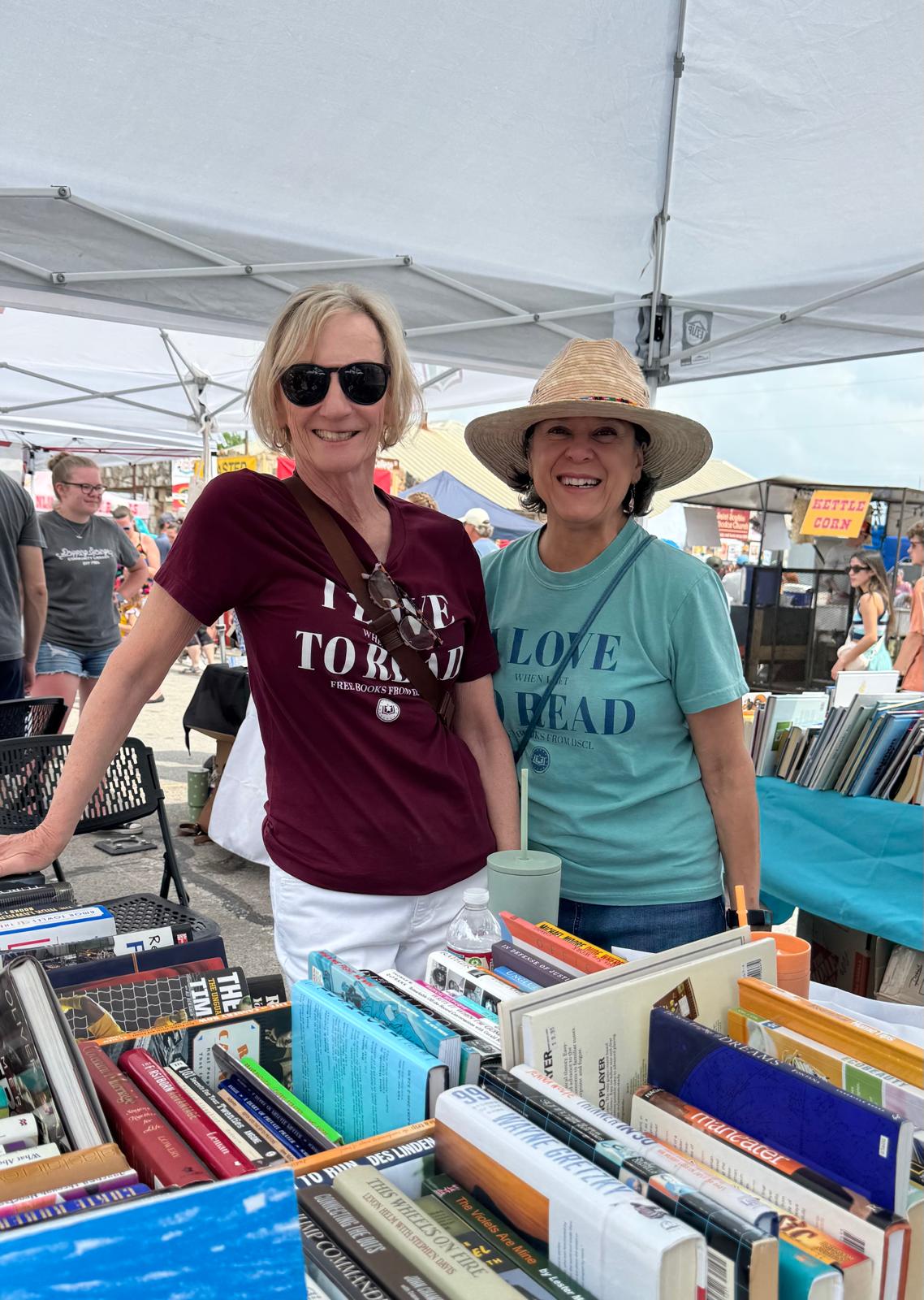 Two volunteers in 'I love to read' shirts behind boxes of books at a Friends book sale